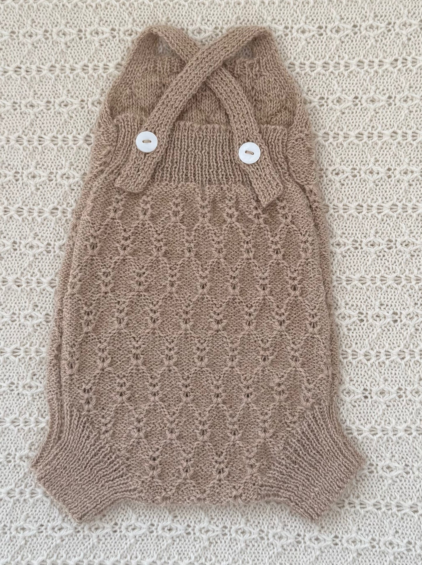 A hand-knit pet sweater in a warm taupe-brown color, featuring a delicate openwork pattern, a halter-style collar with two crossed straps, and two white decorative buttons. The sweater is laid flat on a textured cream lace background.