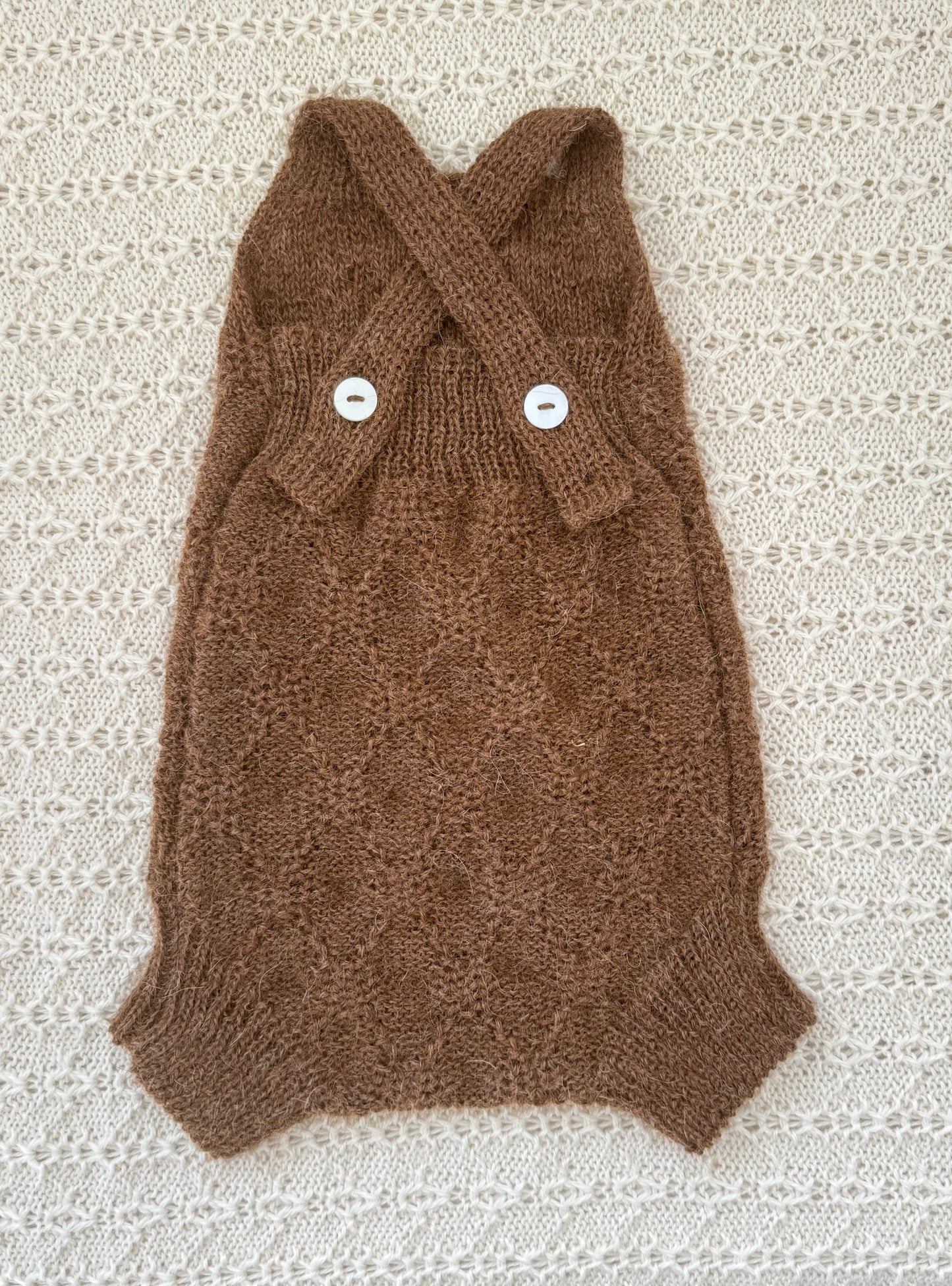 A chocolate alpaca knit baby romper with a crossed straps and two white buttons, laid flat on a textured cream lace background.