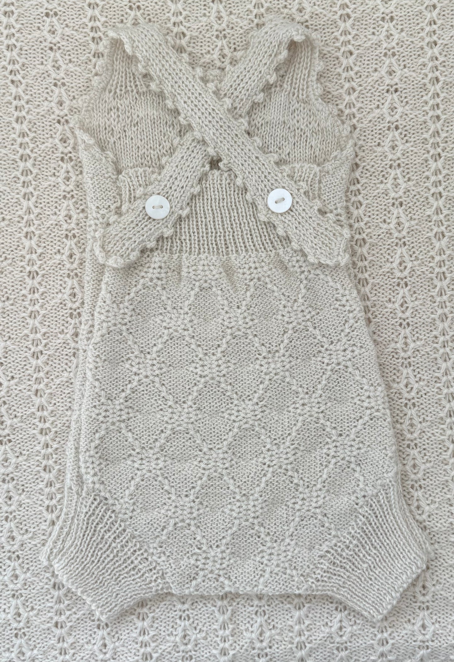 A cream-colored hand-knit baby romper with a diamond lattice pattern, featuring crossed shoulder straps with scalloped edges and two white buttons, laid flat on a matching textured knit blanket.
