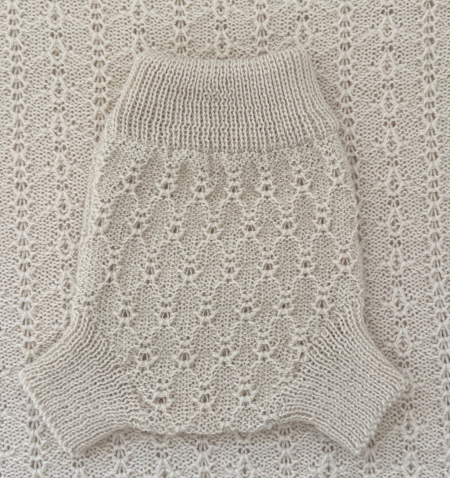 A cream-colored knit pet sweater with a textured openwork pattern and ribbed collar, laid flat on a matching cream knit blanket.