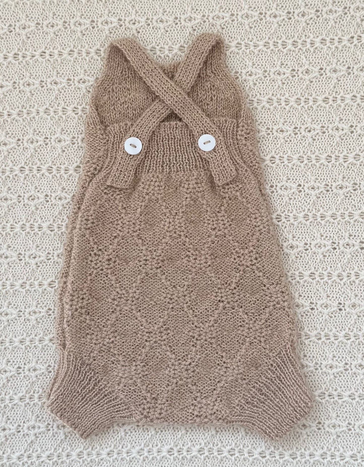 A handmade knit baby romper in a warm taupe-brown color, featuring a diamond-knit pattern, crossed shoulder straps, and two white decorative buttons, laid flat on a textured cream lace background.