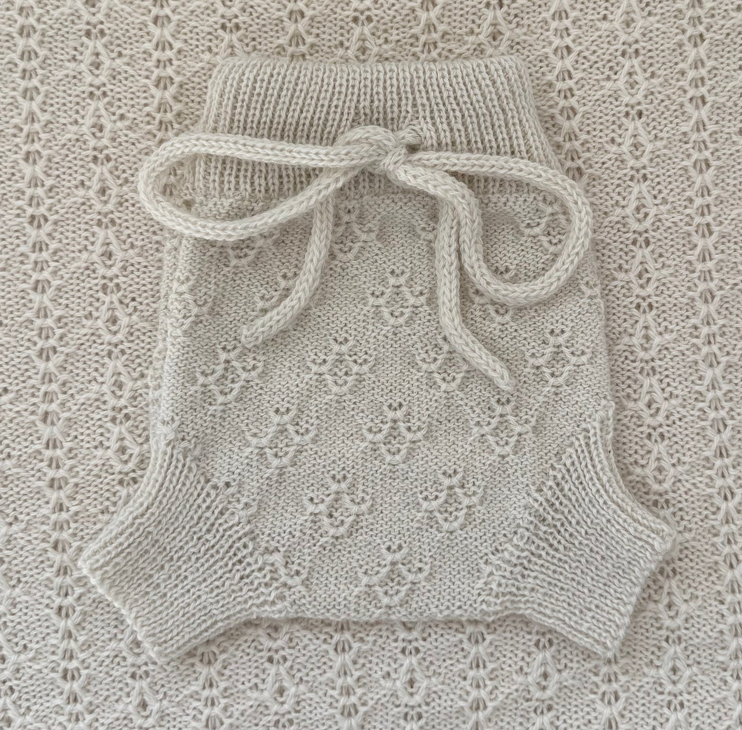 A cream-colored hand-knit baby romper with a lace pattern and a matching knotted bow at the waist, laid flat on a textured lace blanket.