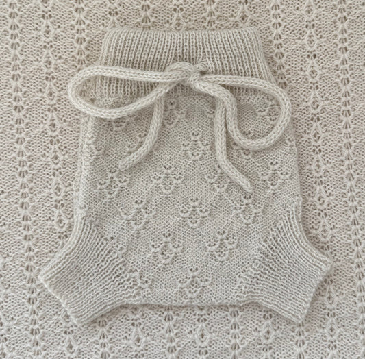 A cream-colored hand-knit baby romper with a lace pattern and a matching knotted bow at the waist, laid flat on a textured lace blanket.