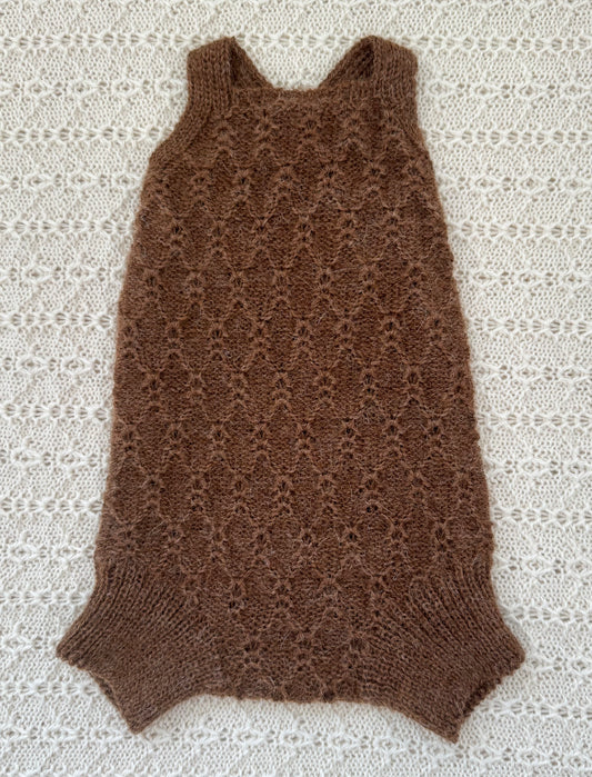 A brown sleeveless knit romper with a textured openwork pattern and ribbed collar, cuffs, and hem, laid flat on a white lace background.
