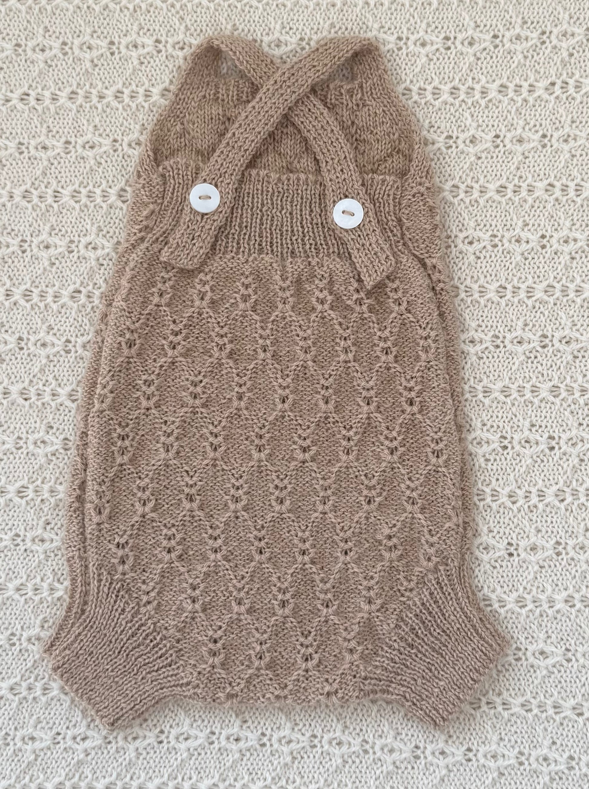 A hand-knit pet sweater in a warm taupe-brown color, featuring a delicate openwork pattern, a halter-style collar with two crossed straps, and two white decorative buttons. The sweater is laid flat on a textured cream lace background.