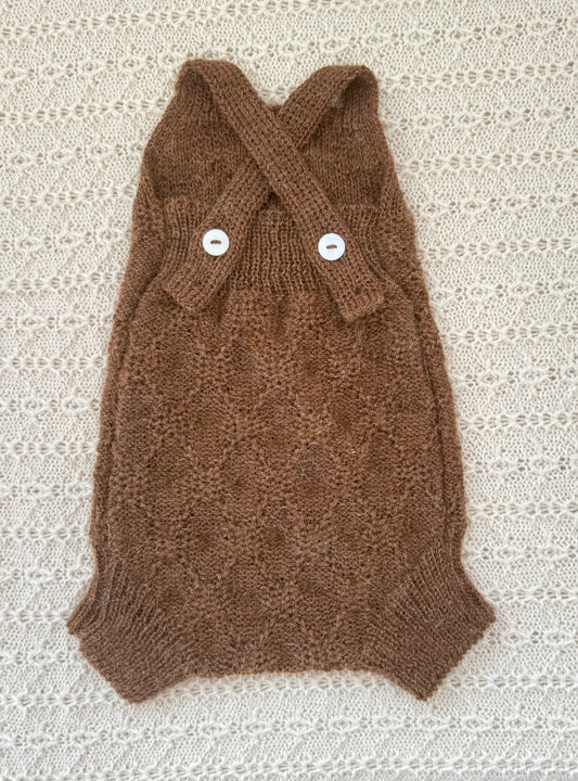 A chocolate alpaca knit baby romper with a crossed straps and two white buttons, laid flat on a textured cream lace background.