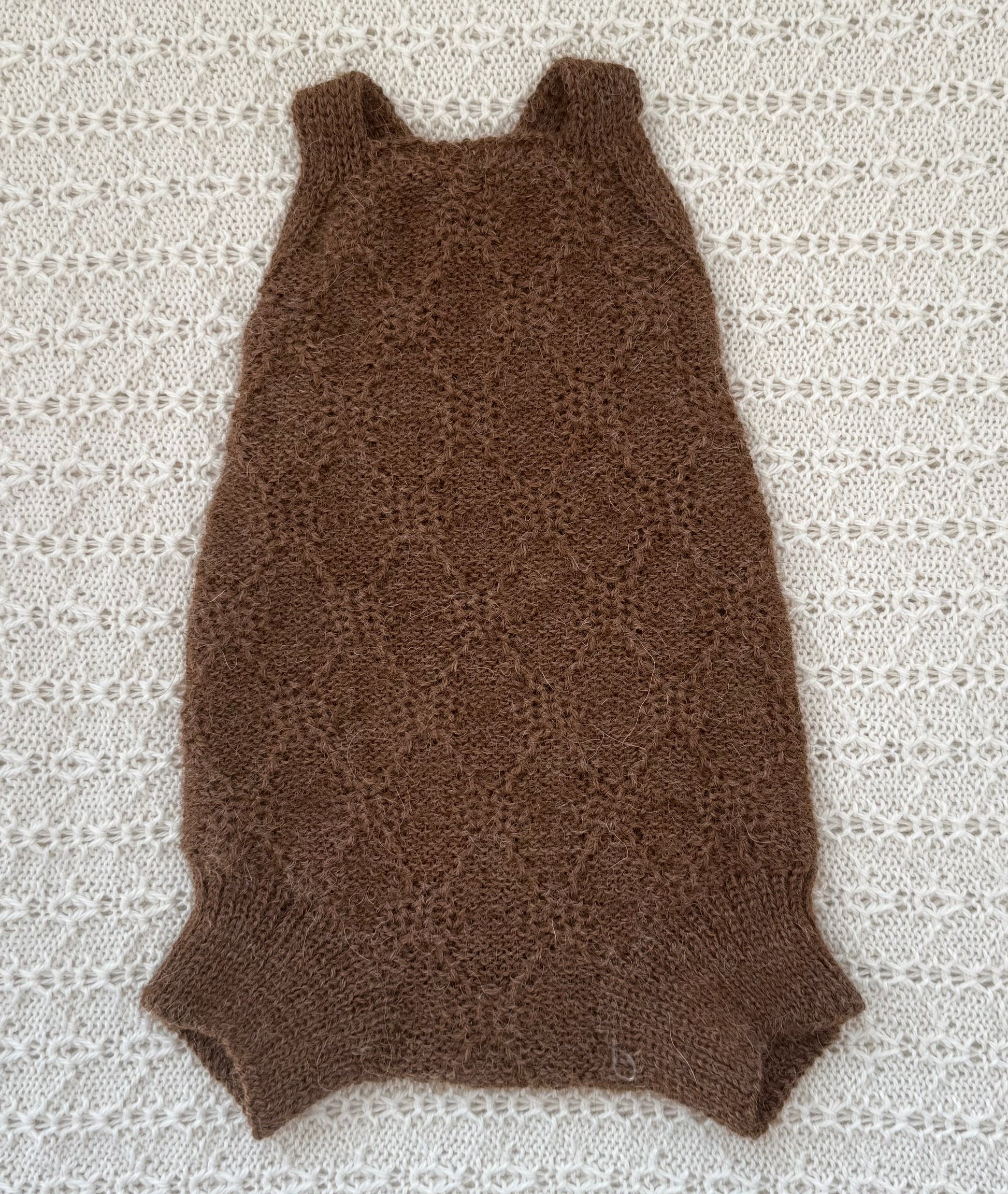 A brown knit sleeveless baby romper with a textured openwork pattern, laid flat on a white lace blanket.
