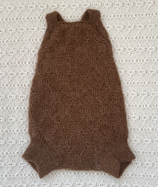 A brown knit sleeveless baby romper with a textured openwork pattern, laid flat on a white lace blanket.