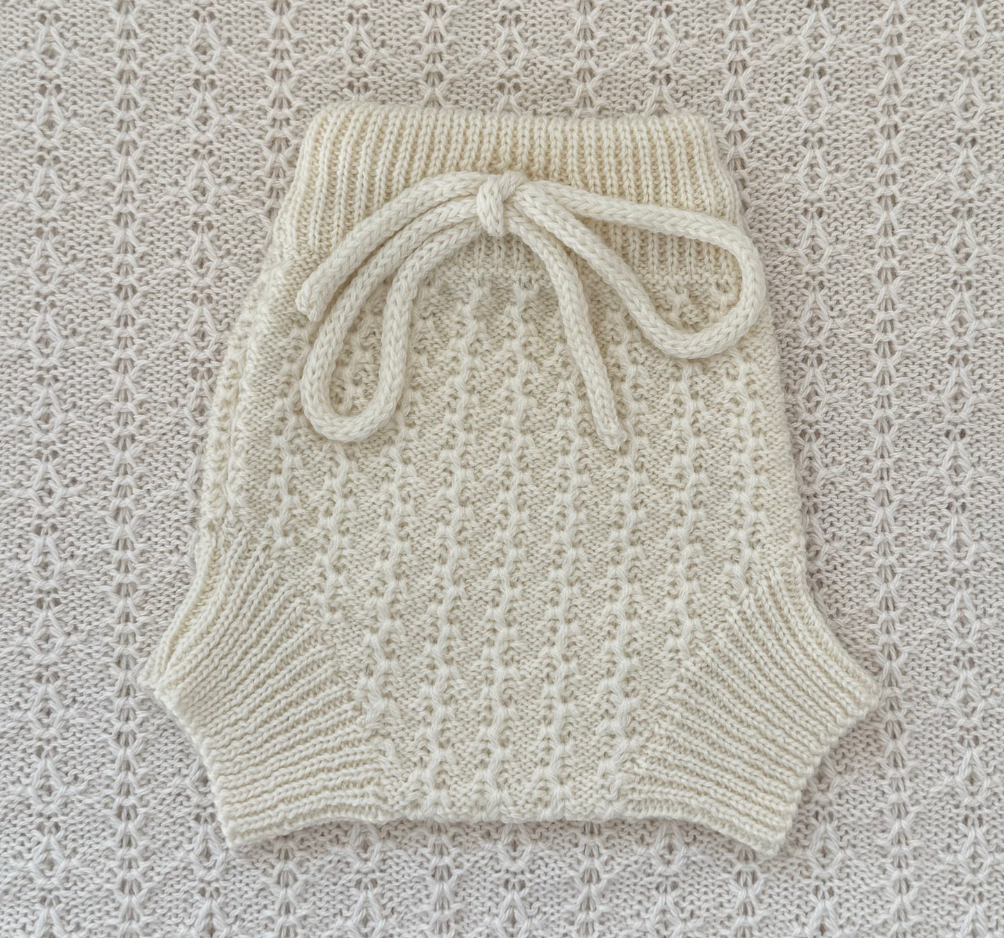 Cream-colored hand-knit baby shorts with a ribbed waistband and a matching knotted bow, laid flat on a textured cream lace background.