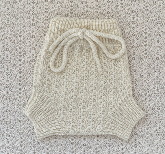 Cream-colored hand-knit baby shorts with a ribbed waistband and a matching knotted bow, laid flat on a textured cream lace background.