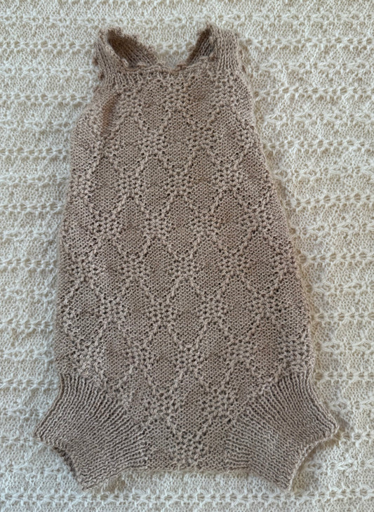A sleeveless, open-knit baby romper in a warm taupe-brown color, laid flat on a textured cream lace blanket.