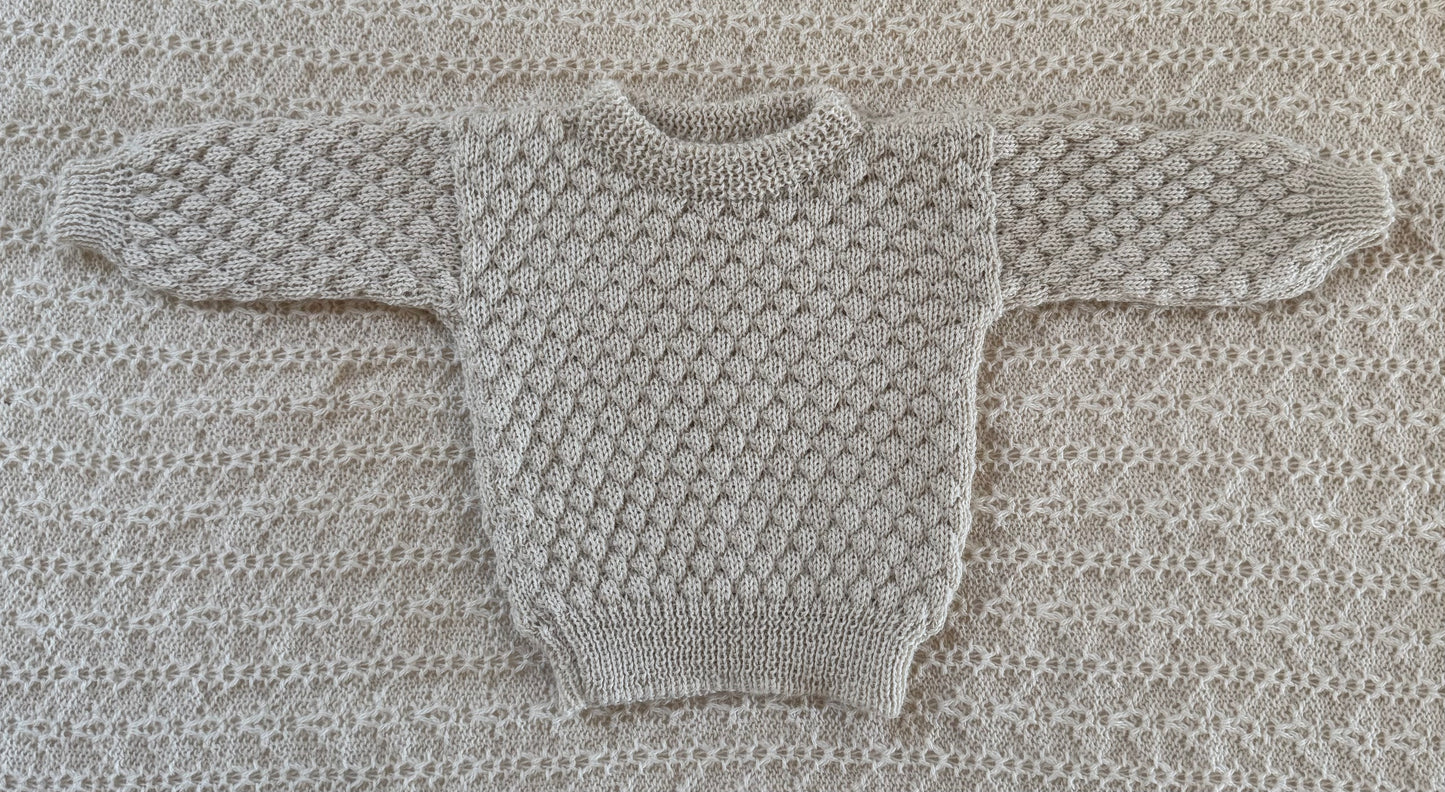 A ivory -knit baby sweater laid flat on a matching textured knit blanket background.