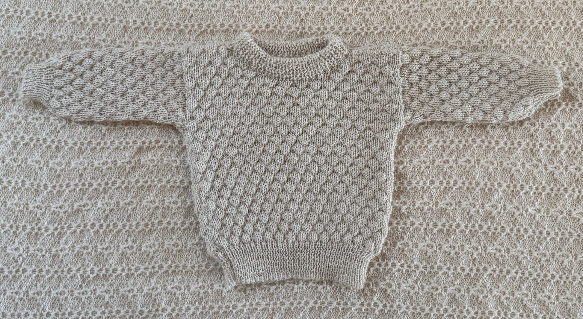A ivory -knit baby sweater laid flat on a matching textured knit blanket background.