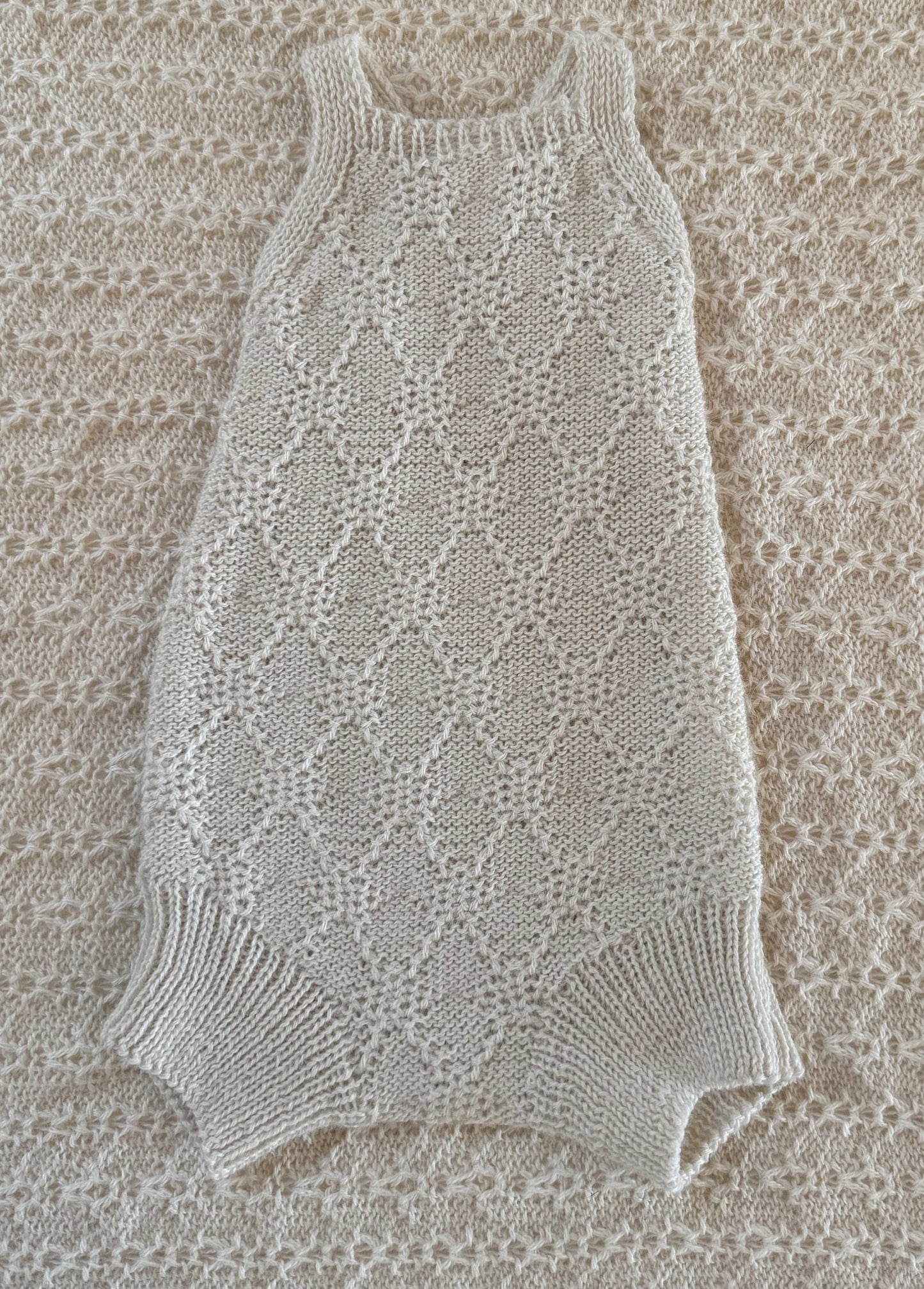 A cream-colored, hand-knit sleeveless baby romper with a textured openwork pattern and ribbed trim, laid flat on a matching cream knit blanket.