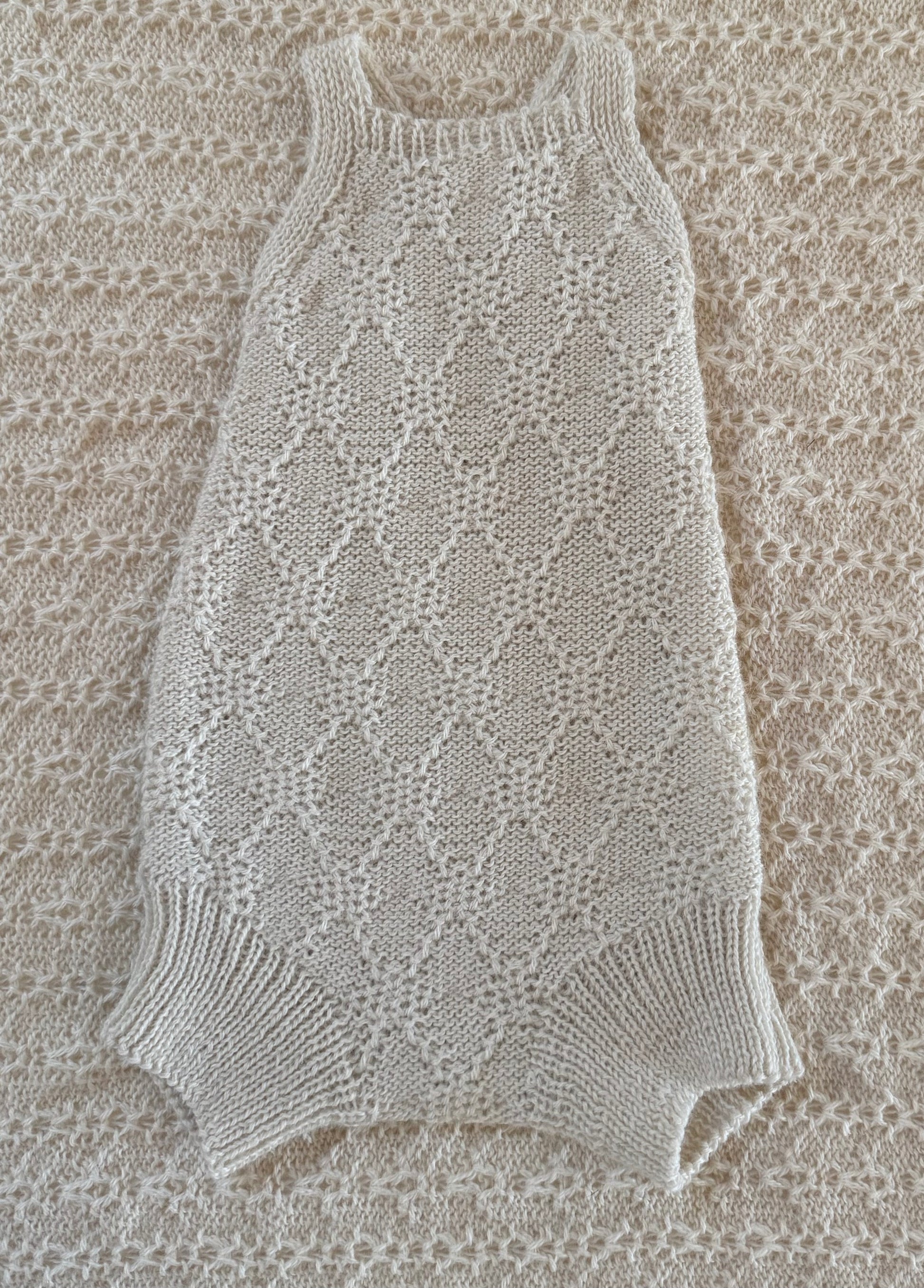 A cream-colored, hand-knit sleeveless baby romper with a textured openwork pattern and ribbed trim, laid flat on a matching cream knit blanket.