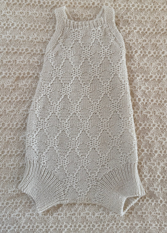 A cream-colored, hand-knit sleeveless baby romper with a textured openwork pattern and ribbed trim, laid flat on a matching cream knit blanket.