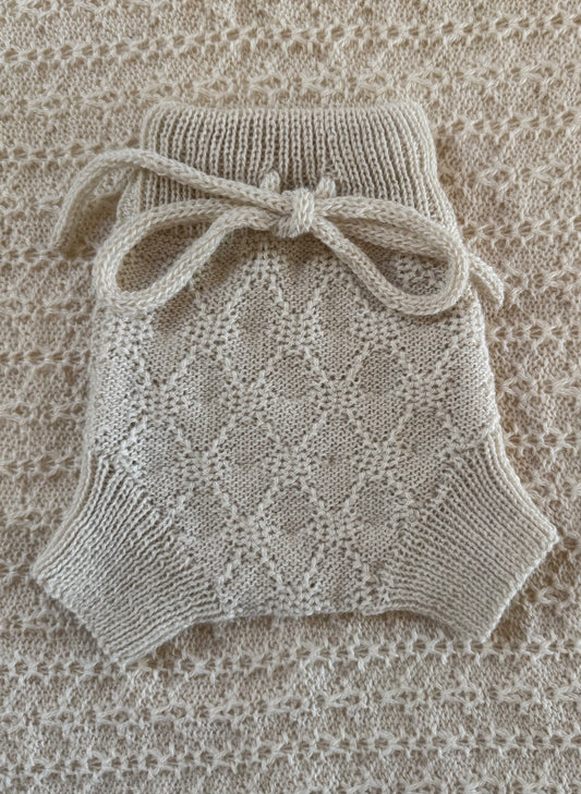 A ivory hand-knit baby bloomers with a textured diamond pattern and a matching knotted bow on the waist, laid flat on a matching knit blanket.