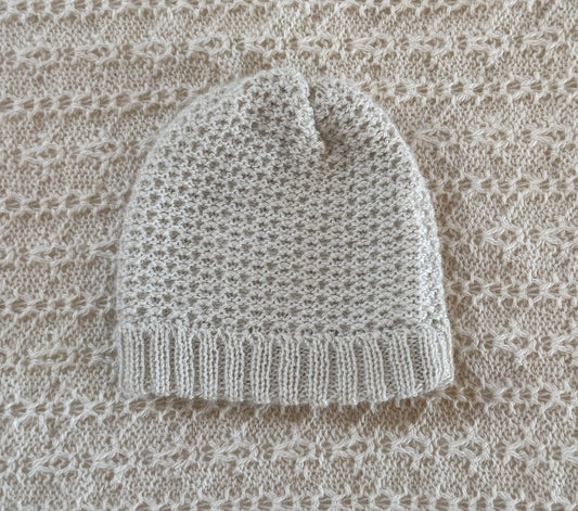 A ivory knit beanie hat with a textured openwork pattern and a ribbed cuff, laid flat on a matching beige knit blanket.