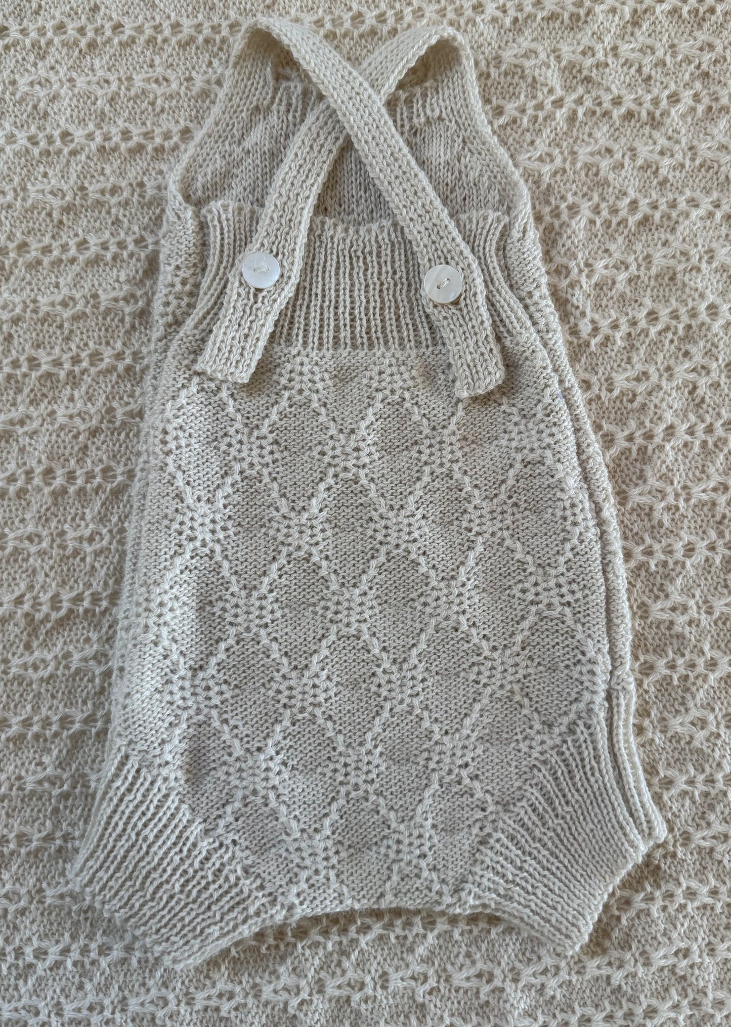 A cream-colored hand-knit baby romper with a diamond lattice pattern, featuring crossed shoulder straps with two decorative buttons, laid flat on a matching textured knit blanket.