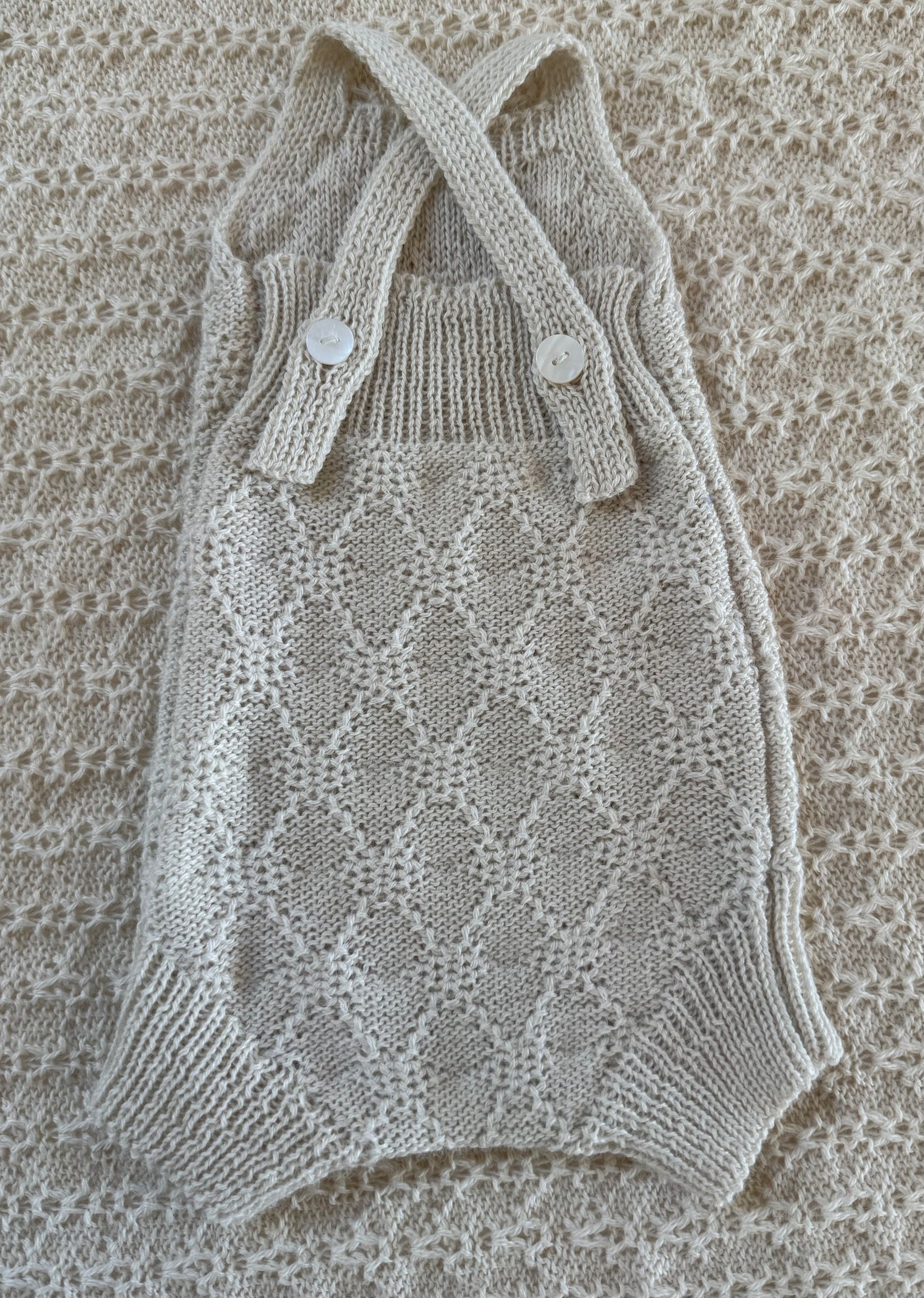 A cream-colored hand-knit baby romper with a diamond lattice pattern, featuring crossed shoulder straps with two decorative buttons, laid flat on a matching textured knit blanket.