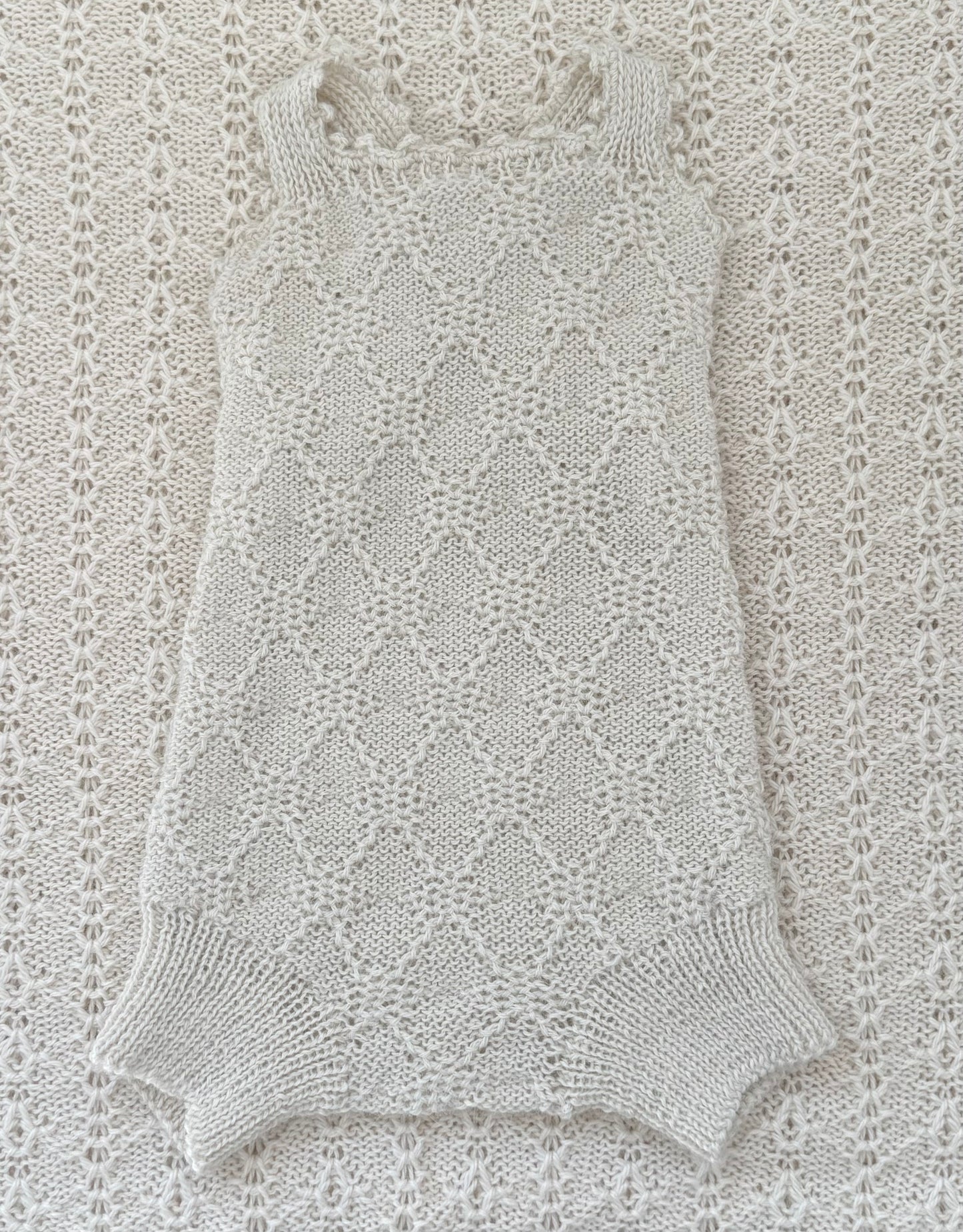 A sleeveless knit romper or bodysuit in a light cream/off-white color, laid flat on a matching textured knit blanket. The garment features a delicate openwork diamond lattice pattern and ribbed trim at the neckline and leg openings.