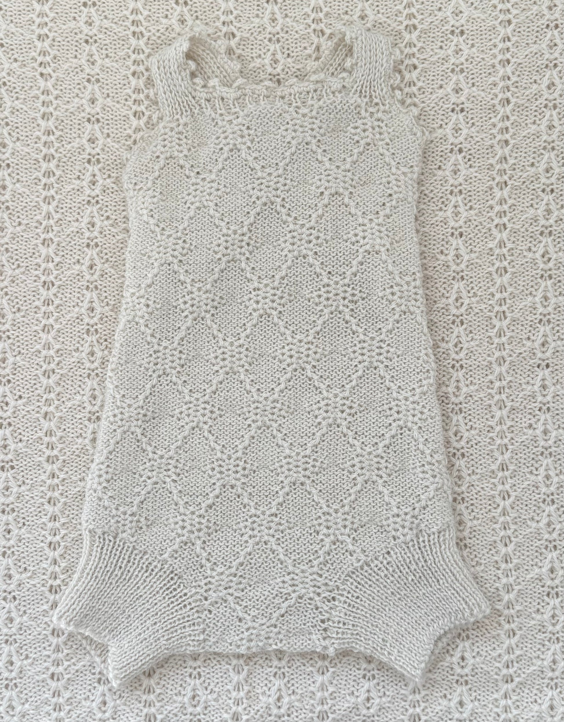 A sleeveless knit romper or bodysuit in a light cream/off-white color, laid flat on a matching textured knit blanket. The garment features a delicate openwork diamond lattice pattern and ribbed trim at the neckline and leg openings.