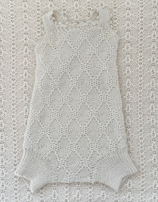 A sleeveless knit romper or bodysuit in a light cream/off-white color, laid flat on a matching textured knit blanket. The garment features a delicate openwork diamond lattice pattern and ribbed trim at the neckline and leg openings.