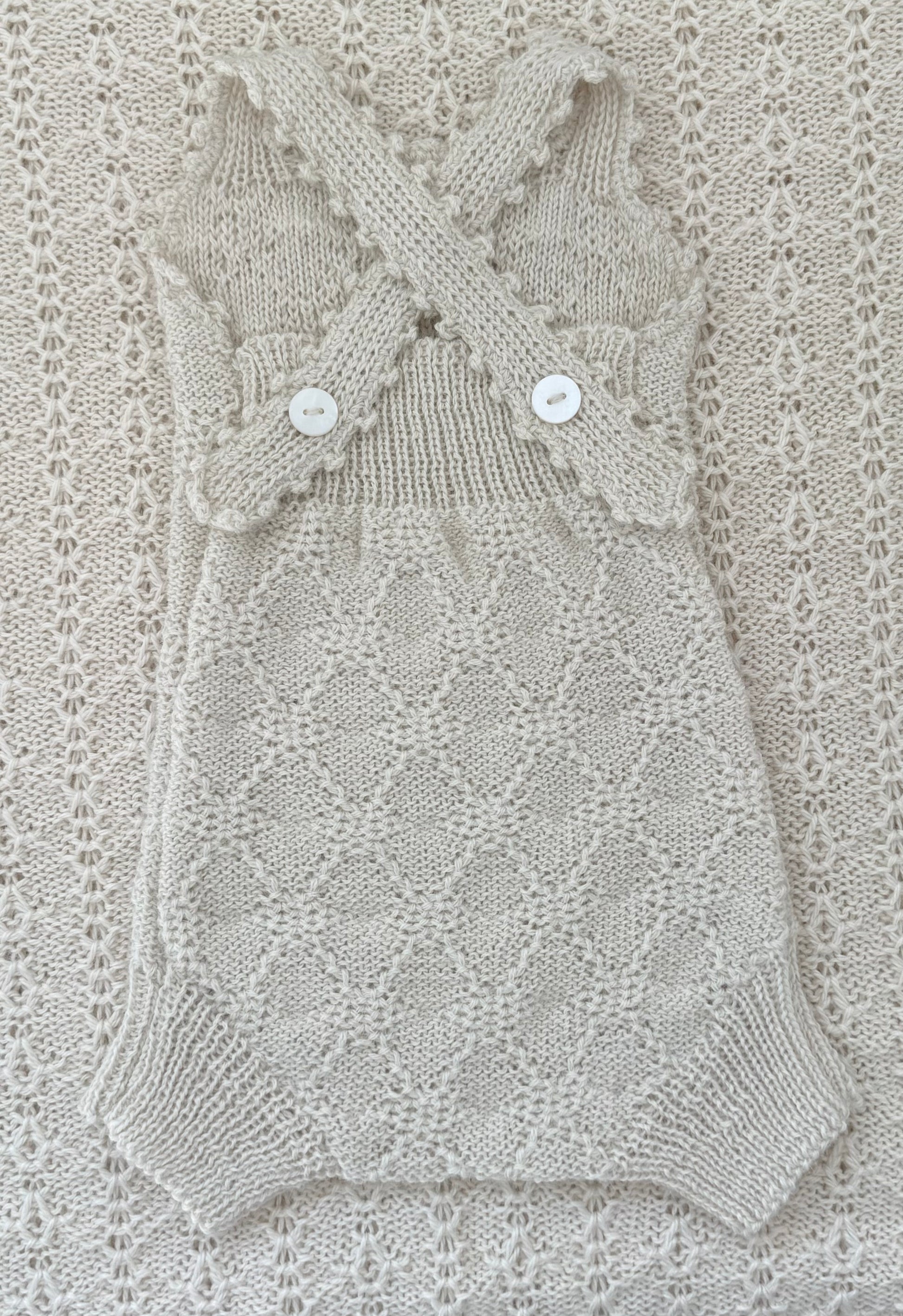 A cream-colored hand-knit baby romper with a diamond lattice pattern, featuring crossed shoulder straps with scalloped edges and two white buttons, laid flat on a matching textured knit blanket.