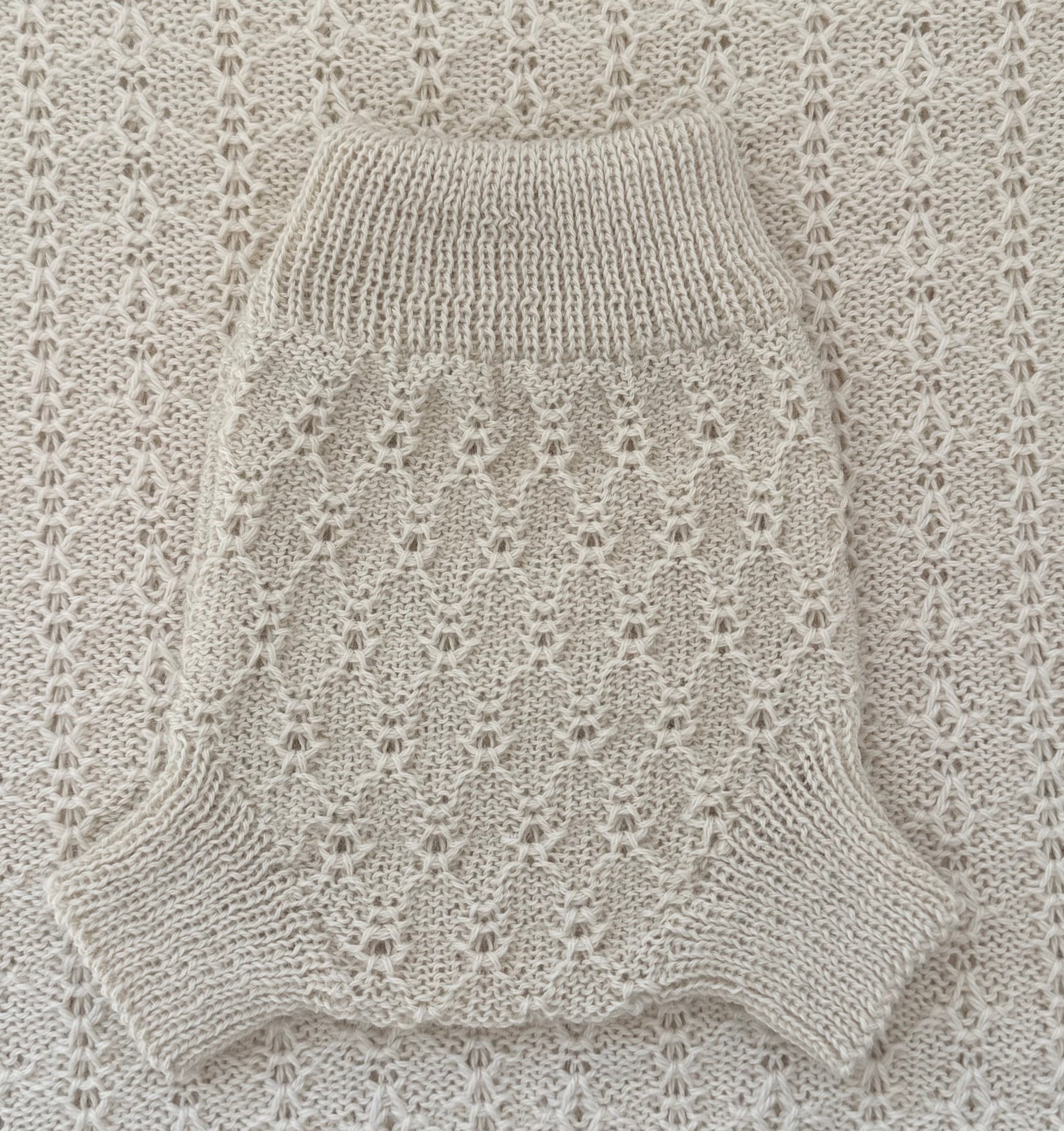 A cream-colored knit pet sweater with a textured openwork pattern and ribbed collar, laid flat on a matching cream knit blanket.
