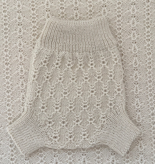 A cream-colored knit pet sweater with a textured openwork pattern and ribbed collar, laid flat on a matching cream knit blanket.