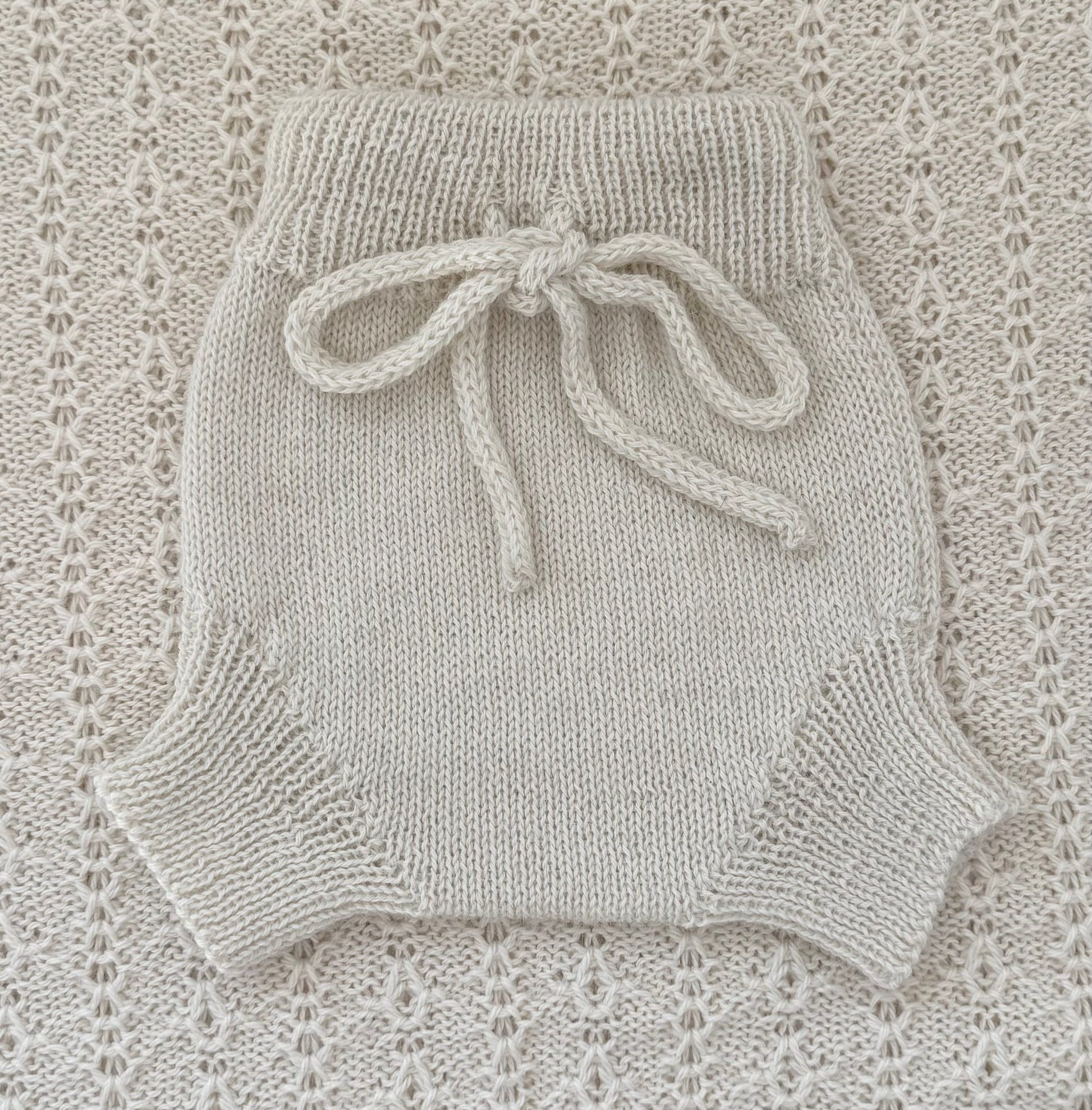 A cream-colored hand-knit baby romper with a matching knotted drawstring waist, laid flat on a textured cream lace blanket.