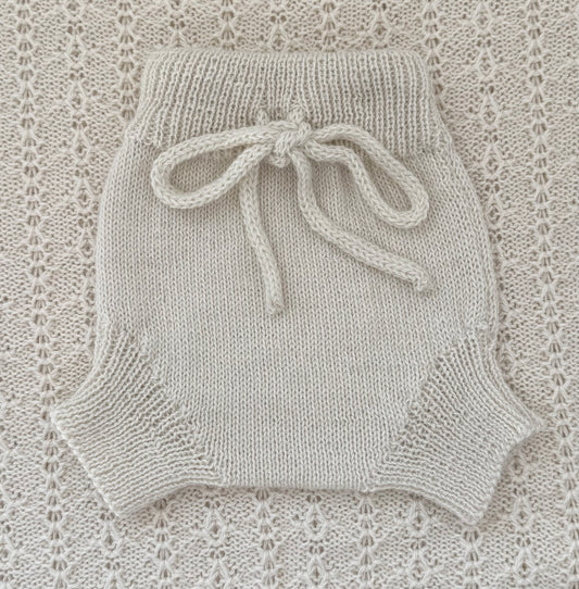 A cream-colored hand-knit baby romper with a matching knotted drawstring waist, laid flat on a textured cream lace blanket.