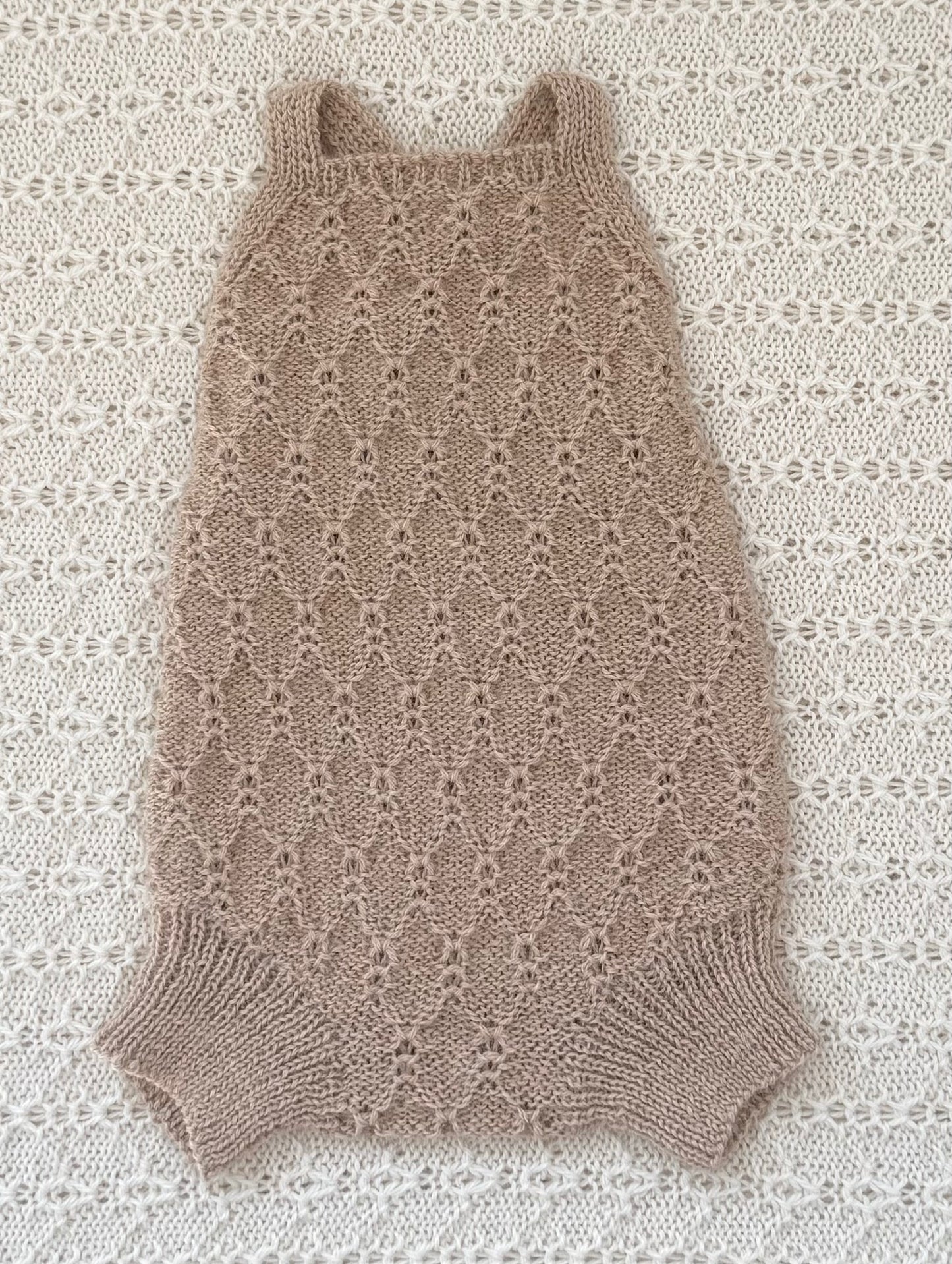 A sleeveless, open-knit baby romper in a warm taupe-beige color, laid flat on a textured cream lace background.