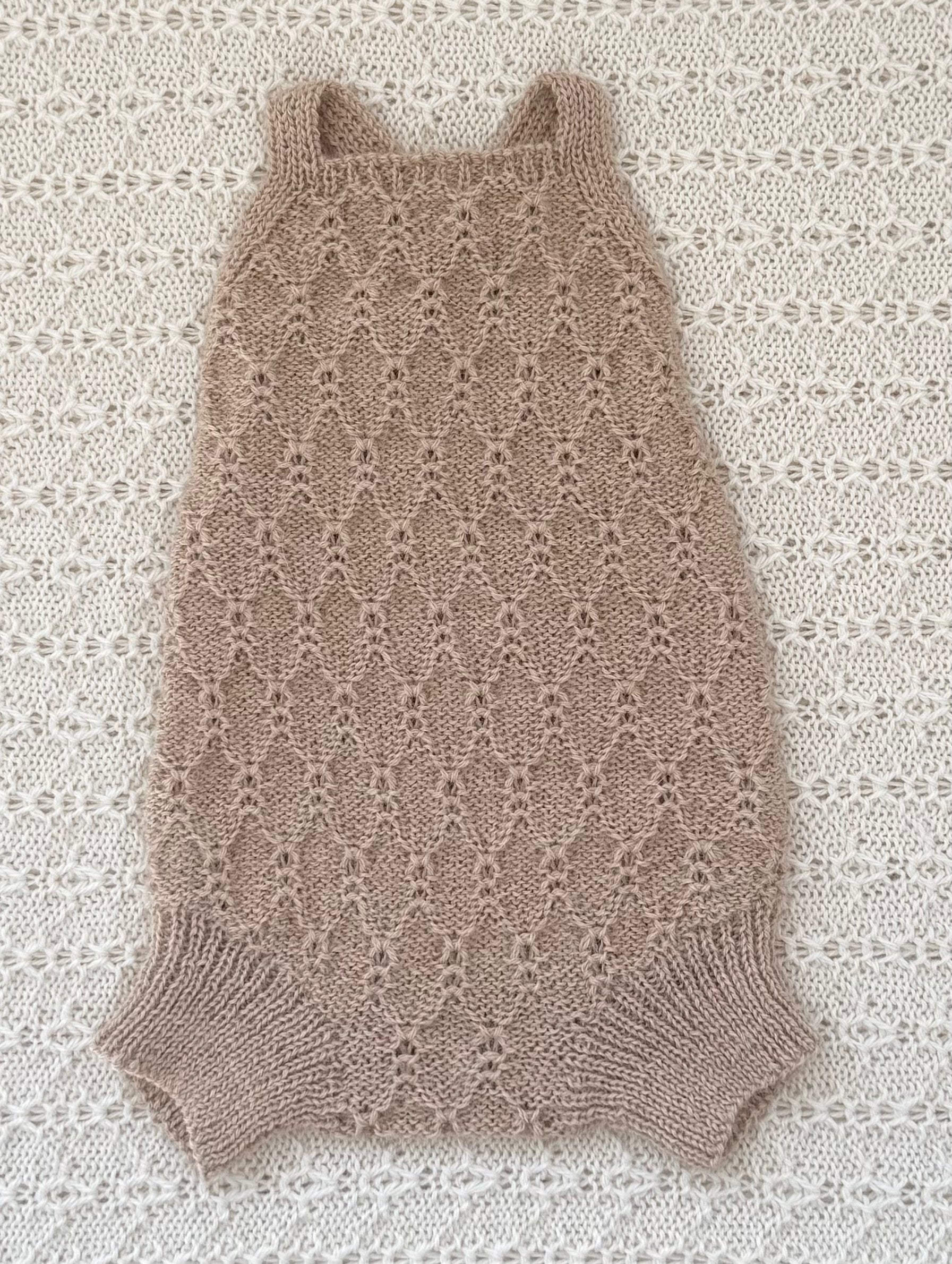 A sleeveless, open-knit baby romper in a warm taupe-beige color, laid flat on a textured cream lace background.