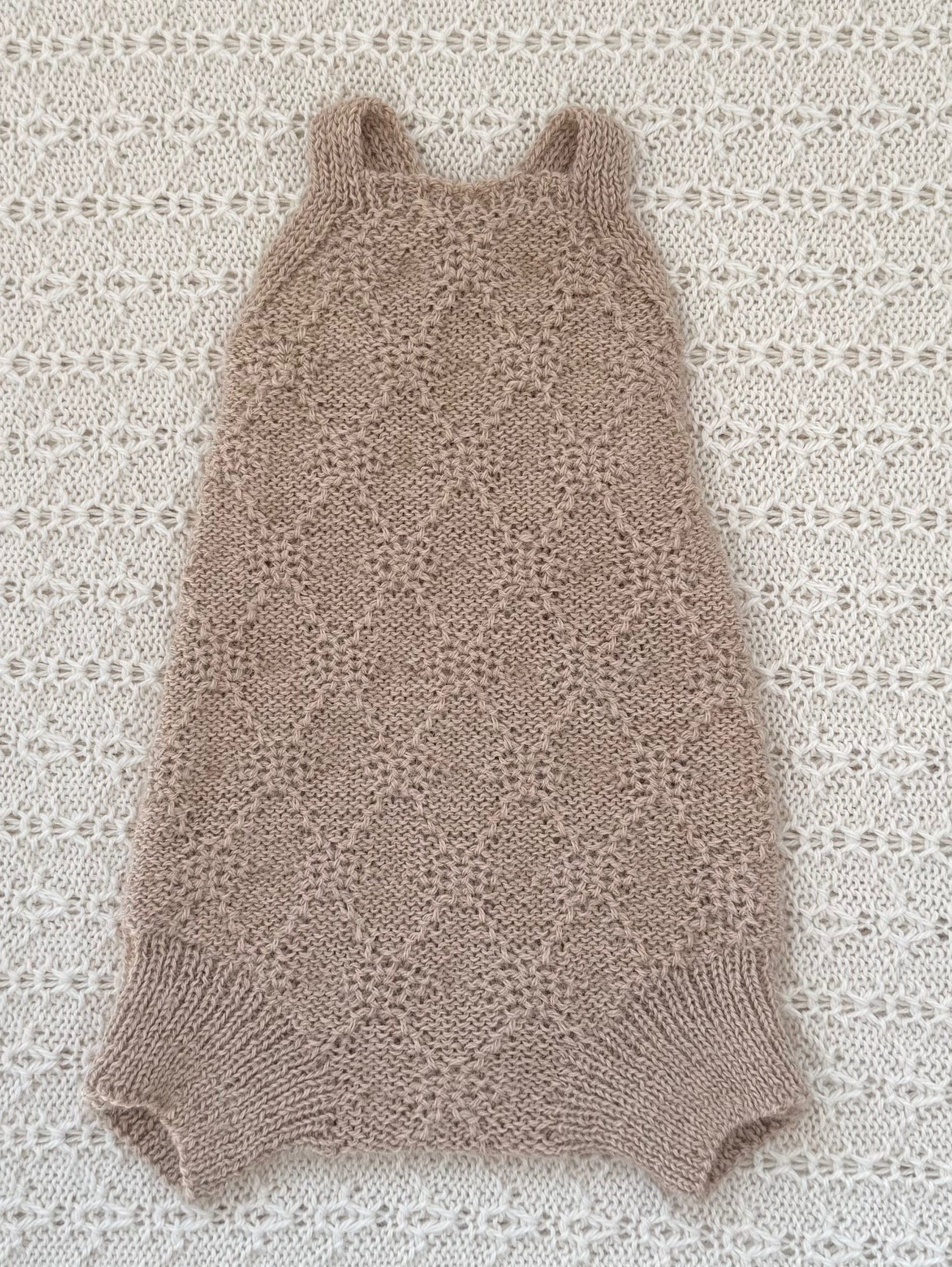 A sleeveless knit baby romper in a light taupe-beige color, laid flat on a textured white crochet blanket.