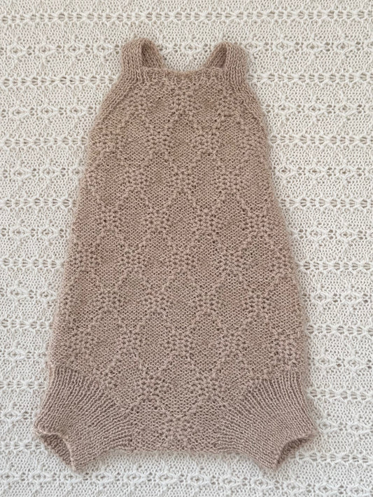 A sleeveless knit baby romper in a light taupe-beige color, laid flat on a textured white crochet blanket.