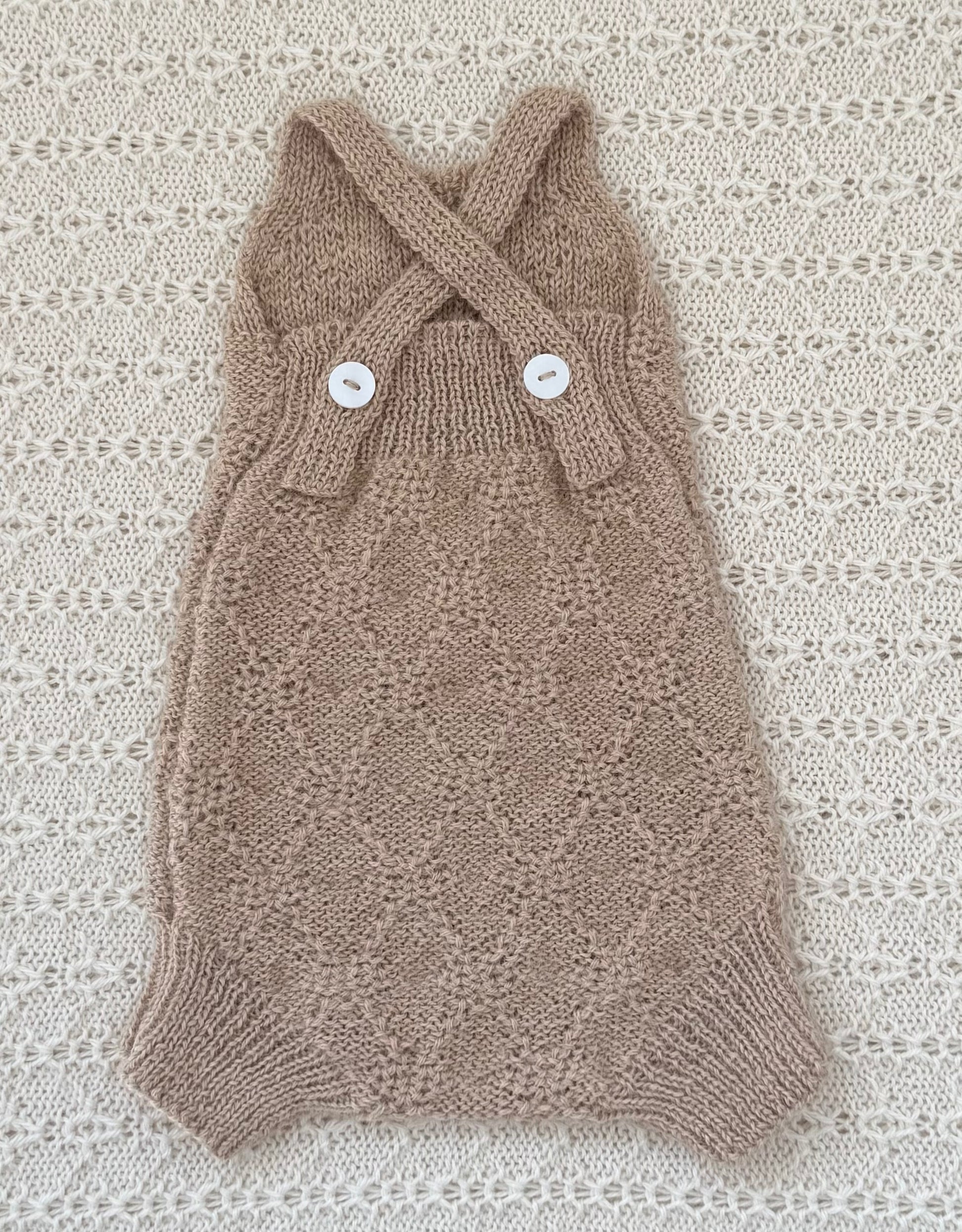A handmade knit baby romper in a warm taupe-brown color, featuring a diamond-knit pattern, crossed shoulder straps, and two white decorative buttons, laid flat on a textured cream lace background.